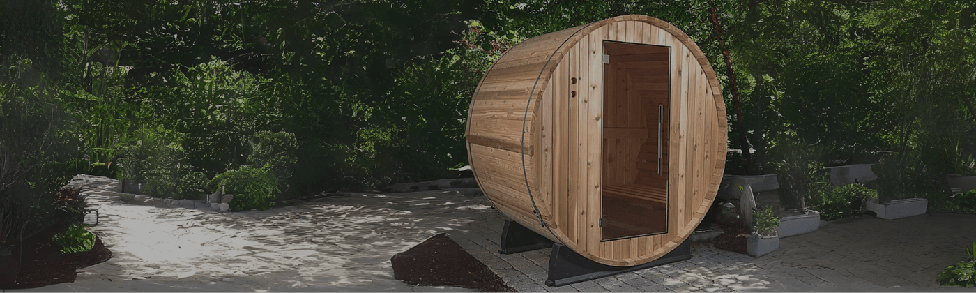 An Almost Heaven outdoor wooden barrel sauna with a glass door sitting on a stone patio surrounded by lush green trees.