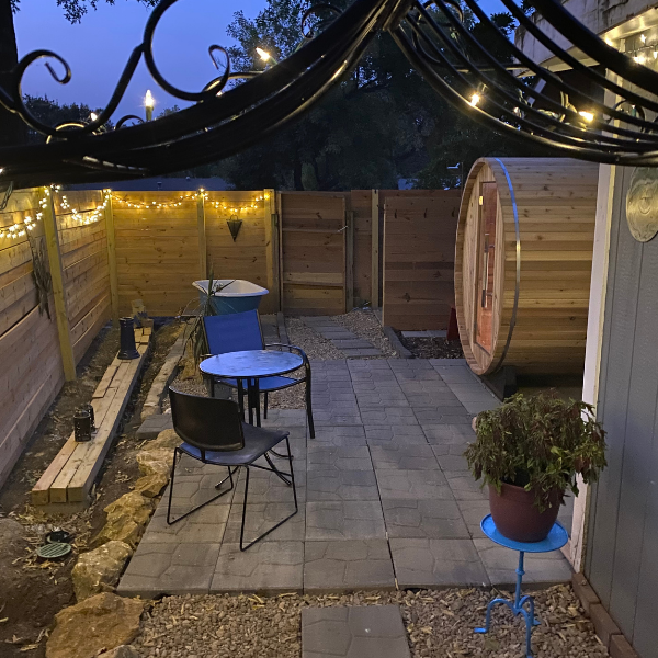 An outdoor wooden barrel sauna situated in a paved backyard courtyard at dusk, featuring a patio table, chairs, and warm string lights along the fence.
