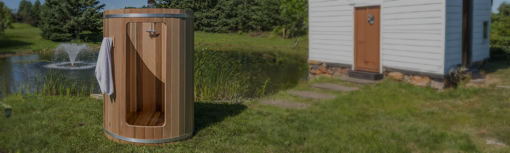 A Dundalk outdoor wooden barrel shower stall with a towel hanging on the side, located on a grassy lawn near a pond and a small house.