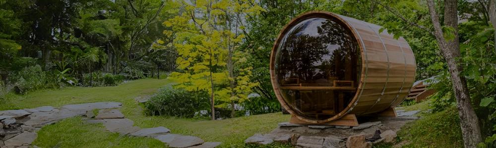 A Dundalk wooden barrel sauna featuring a full panoramic acrylic glass dome back window, positioned outdoors on a grassy hill surrounded by trees.