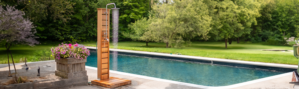 A wooden panel outdoor shower running with water next to a backyard swimming pool surrounded by a green lawn.