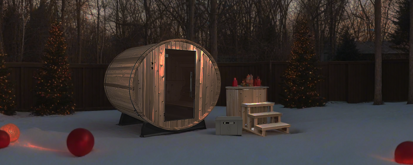 A wooden barrel sauna and matching cold plunge tub in a snow-covered backyard with festive holiday lights at dusk.