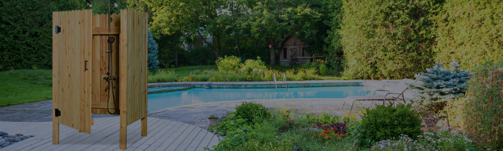 A wooden outdoor shower stall with an open door next to a backyard swimming pool surrounded by trees.