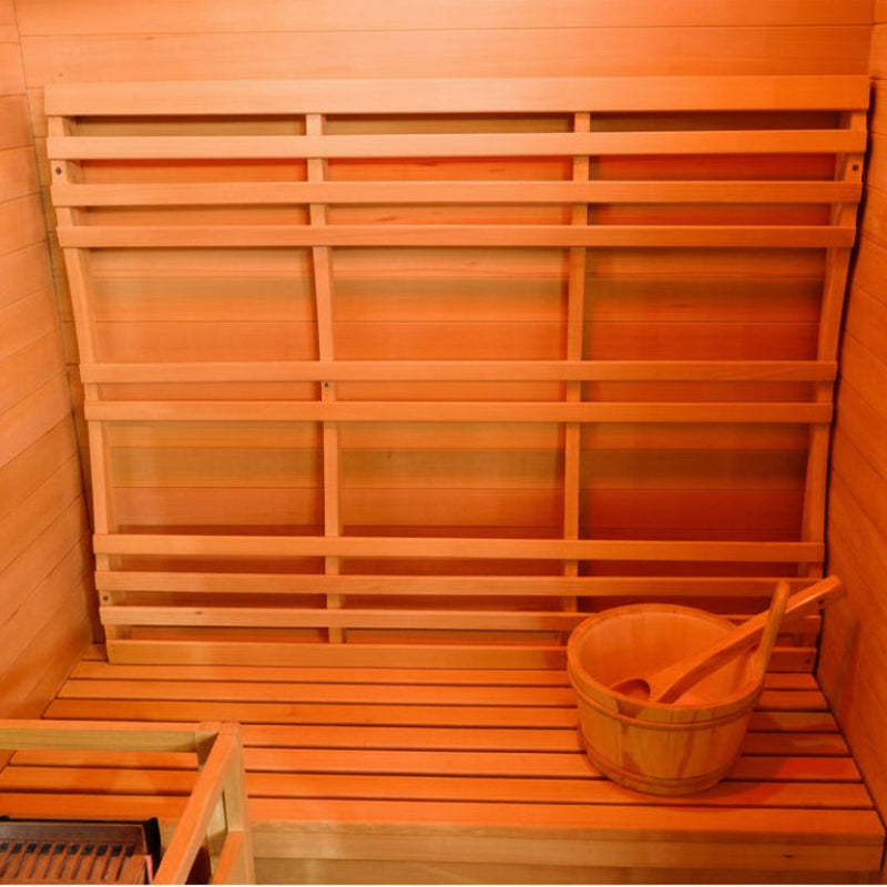 A wooden sauna interior with tiered slatted benches, a hanging wooden bucket, and a wooden paddle inside, shown under warm orange lighting.