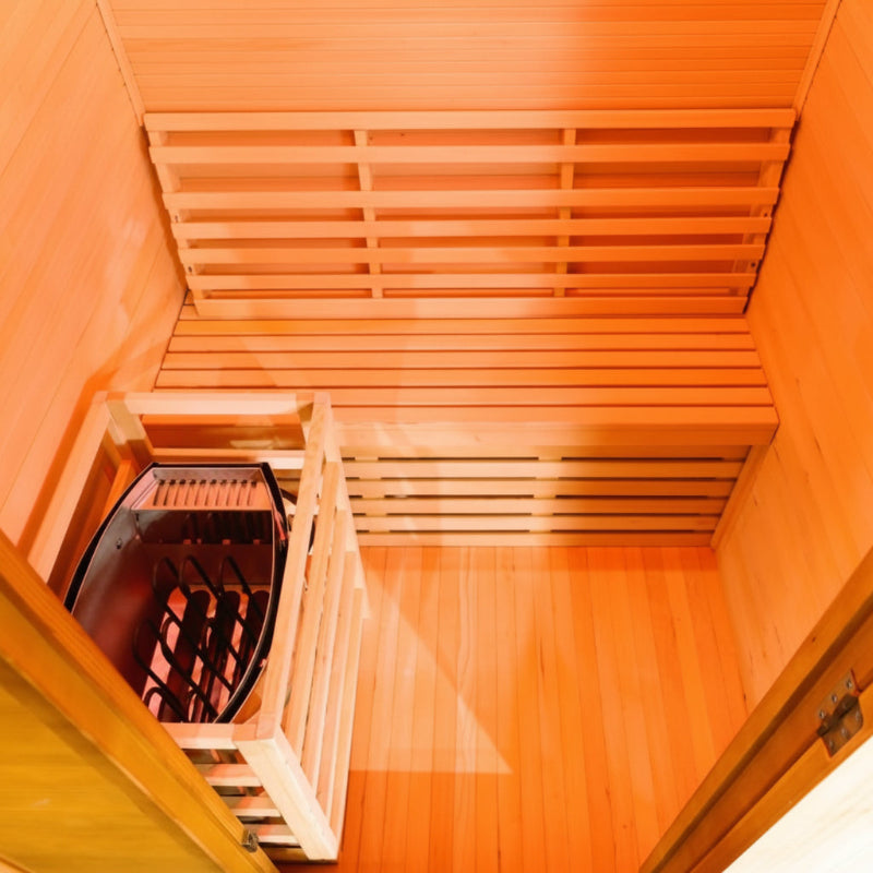 An overhead view of a wooden sauna interior with tiered wooden benches and a dark metal infrared sauna box on the left, shown against a white background.