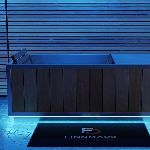 A modern outdoor hot tub with a dark wood-paneled exterior and a matching metal rim, shown under cool blue ambient lighting. A black rectangular mat in front displays the logo and name FINNMARK DESIGNS.