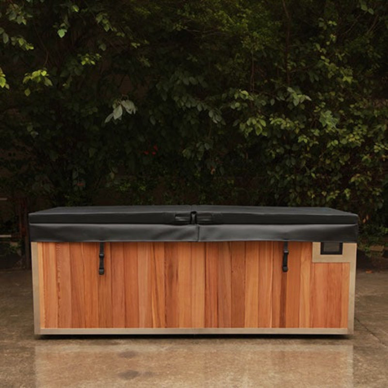 A rectangular outdoor storage chest with a warm brown wood-look front, light beige sides, and a dark gray cover, photographed outdoors against a background of green foliage and a concrete ground.