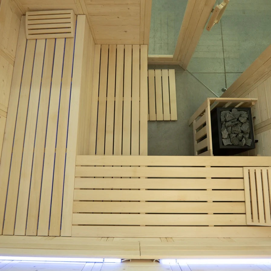 A wooden sauna interior with light natural wood slats, a slatted wall, bench seating, and a black stone firebox filled with dark stones.
