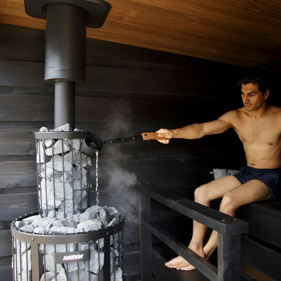 Man sitting in a sauna using a ladle to pour water over rocks in a metal container.