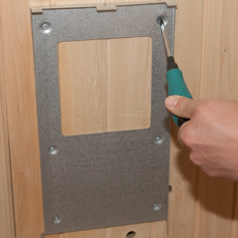 A metal mounting plate with a square cutout is being installed against a light wood surface, with a hand holding a screwdriver.