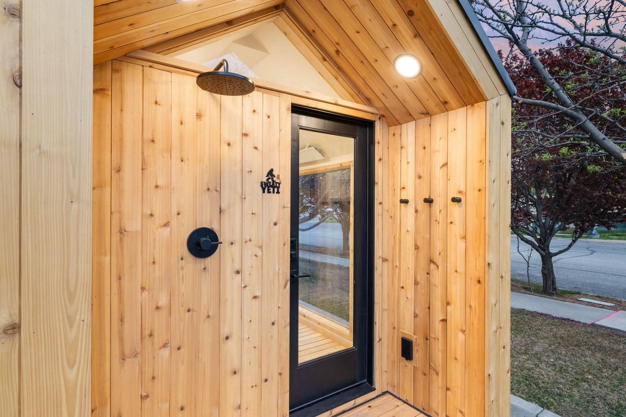 A modern tiny home exterior featuring light natural wood siding, a black-framed glass entry door, a wall-mounted shower head, and a small outdoor seating area on a wooden deck.