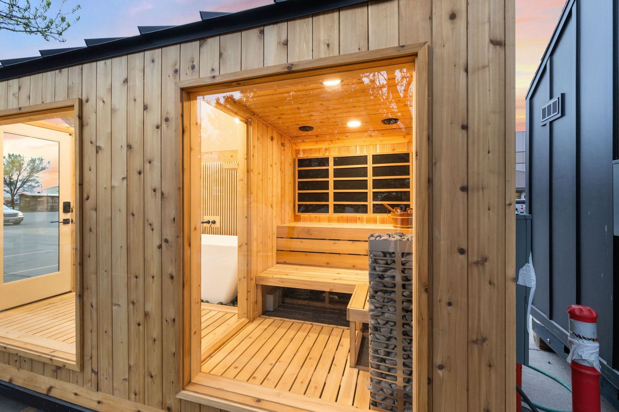A modern wooden sauna cabin with a warm wood-paneled exterior and an open front door revealing a sauna interior with wooden benches, a stone fire column, and a glass window. A glass door is visible on the left, and another dark cabin is partially visible on the right.