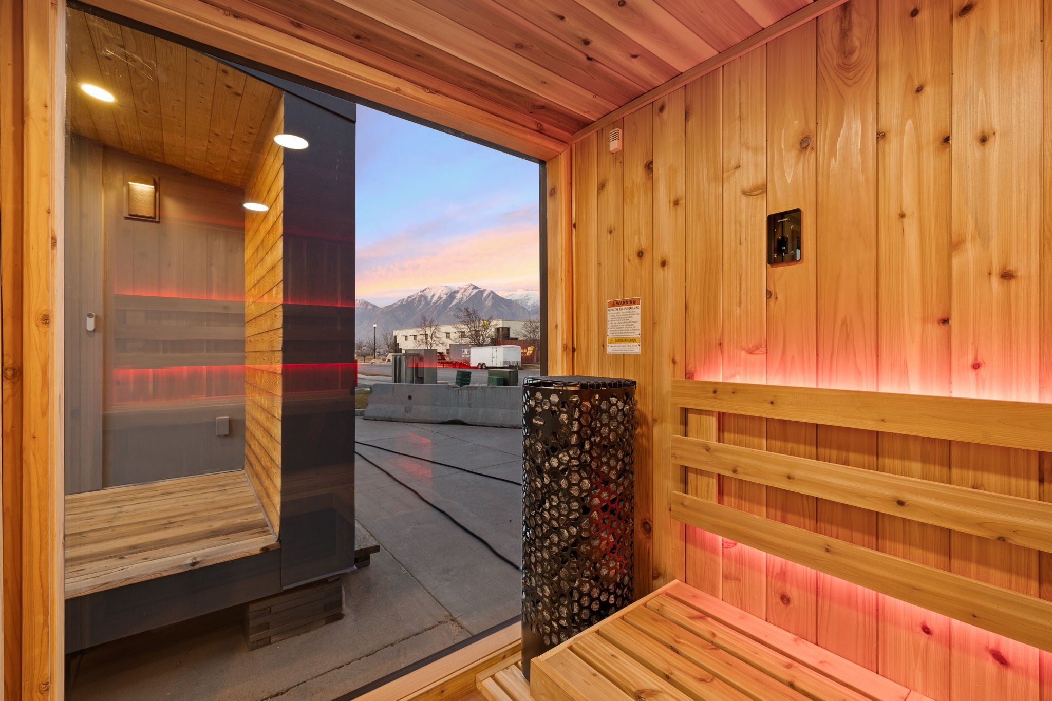Wooden sauna interior with built-in benches, ambient red lighting, and an open door showing an outdoor mountain view at dusk.