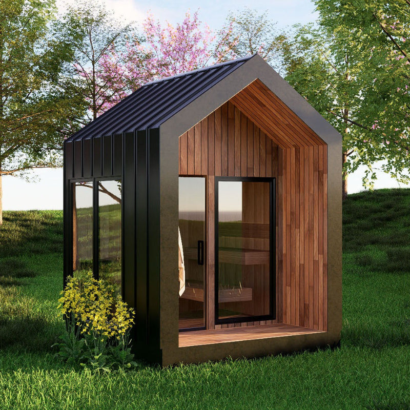 A modern small cabin or sauna with a dark metal exterior, wood-paneled gable front, and large glass doors, shown outdoors on a grassy lawn with trees in the background.