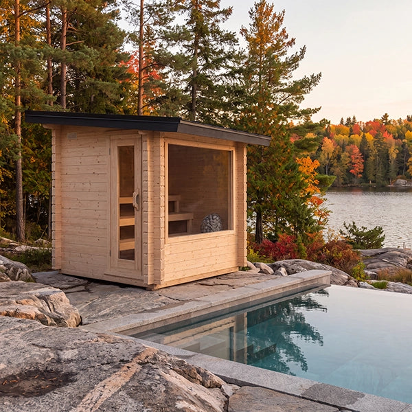 A small light wood cabin sauna with a glass door and large window sits beside a stone pool, surrounded by trees and a lake at sunset.