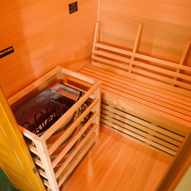A wooden sauna interior with slatted benches and a metal heating panel visible inside the room.
