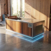 A modern outdoor kitchen island with a wood-paneled front, stainless steel top and trim, and integrated blue LED under-glow lighting, shown in a rustic indoor-outdoor space with brick and wood walls.