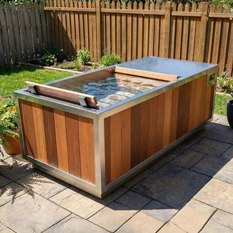A rectangular outdoor hot tub or plunge pool with a wood-look exterior and stainless steel trim, shown on a stone patio in a backyard with a wooden fence and grass.
