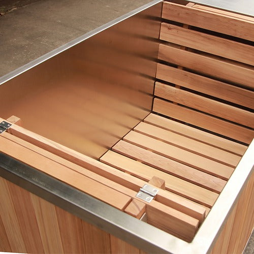 Close-up of a wooden hot tub or sauna interior with light natural wood slats and a brushed metal rim, shown in a warehouse-like setting.