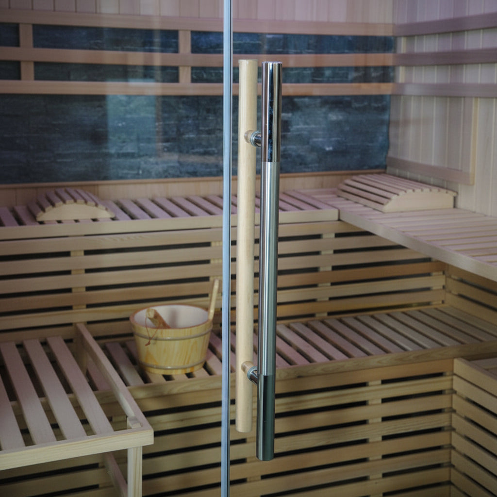A modern glass door handle with a vertical chrome silver handle and a natural wood handle, shown installed on a glass door in front of a wooden sauna interior with a small wooden bucket.