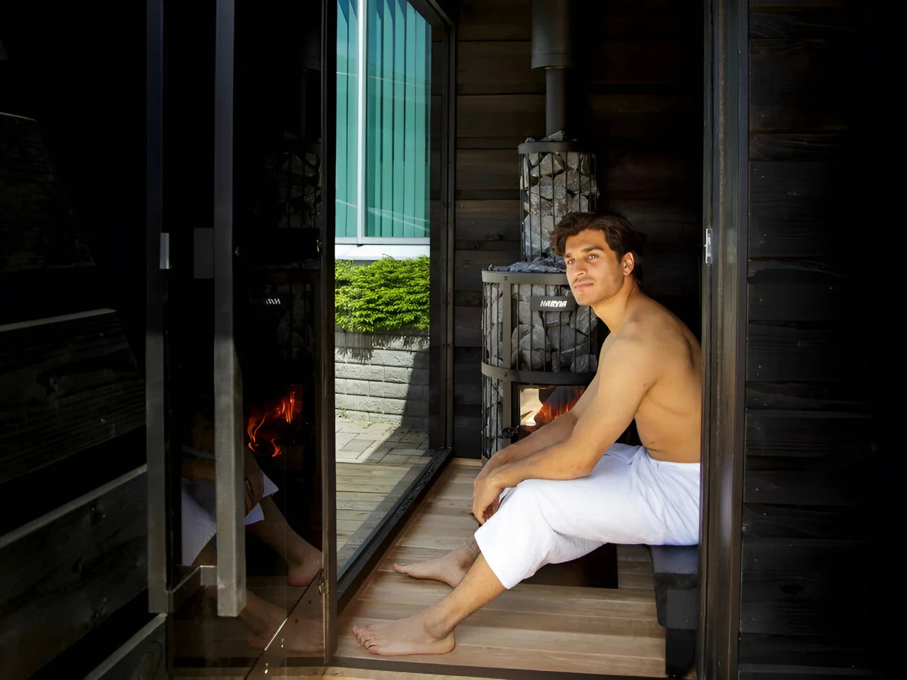 A man sitting inside the sauna with a wood burning heater on the side