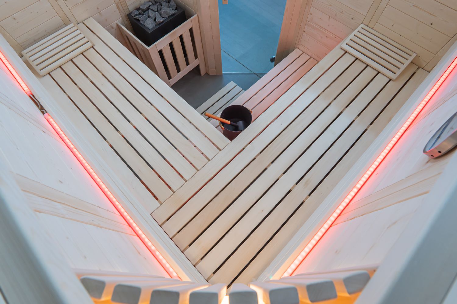 An overhead view of a wooden sauna interior with light natural wood benches, red LED strip lighting along the walls, and a wood fire basket with logs.