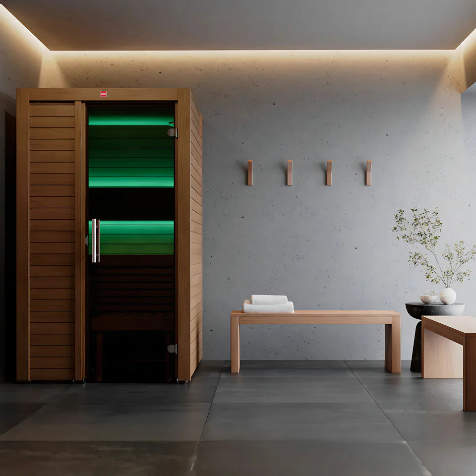 A modern wooden infrared sauna in a minimalist spa-like room with a concrete wall, built-in warm lighting, a bench with folded towels, and a small plant.