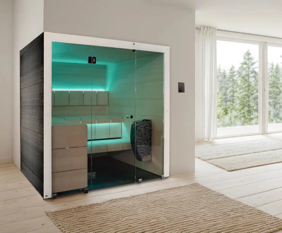 A modern glass sauna cabin with sliding doors, illuminated by bright green LED lighting, set in a minimalist room with light wood floors and a large window showing trees outside.