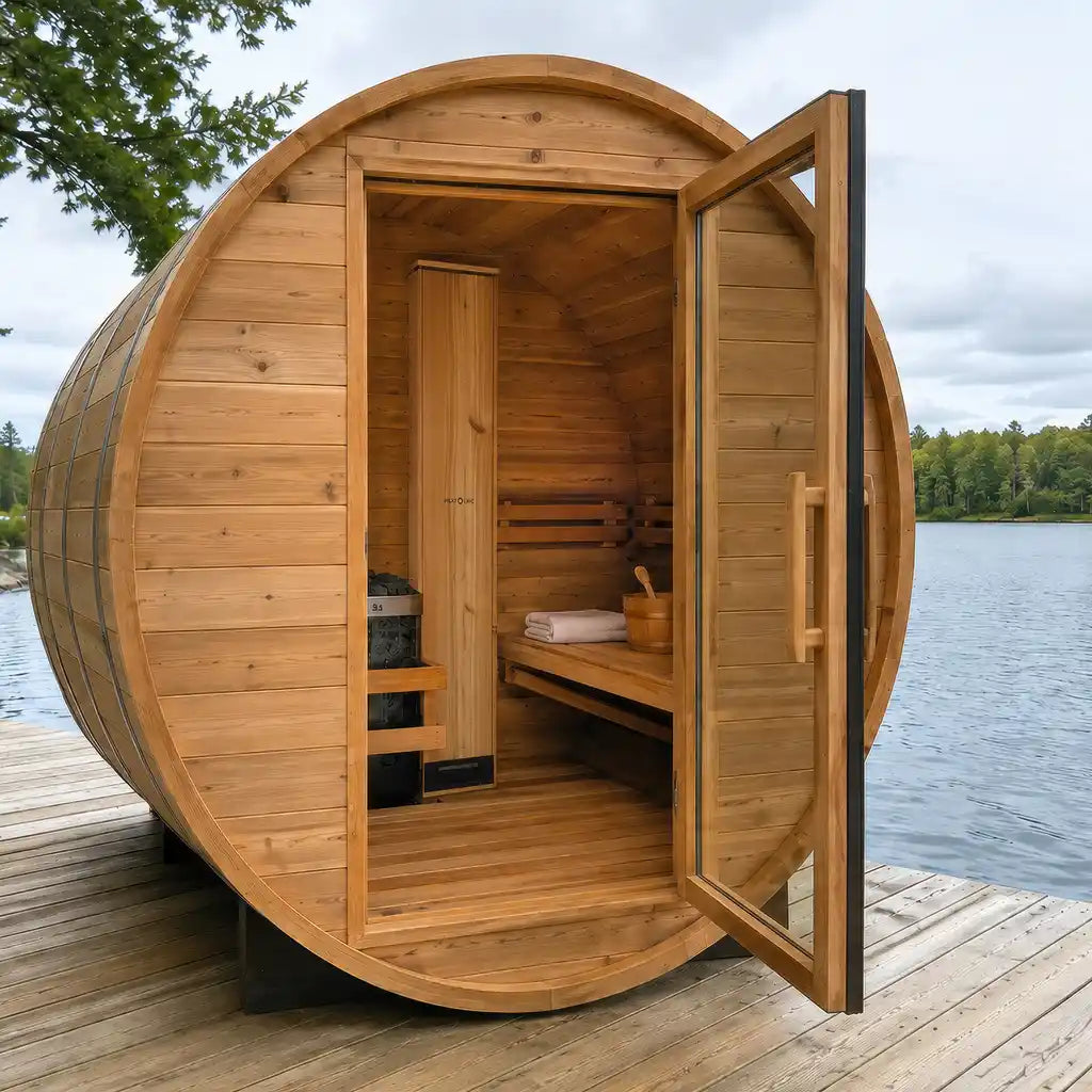 A wooden sauna cabin made from repurposed barrels, sitting on a wooden dock next to a calm lake with trees in the background. The front door is open, revealing a wooden interior with a sauna bench, steam table, and a brazier. CRM Showers Sauna Next Heat Circ