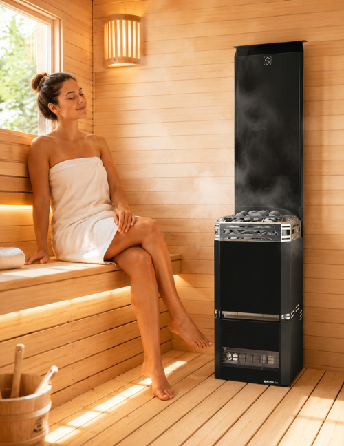 A woman wrapped in a white towel sits on a wooden sauna bench beside a tall black sauna stove filled with hot stones, in a warm wood-paneled sauna room.
