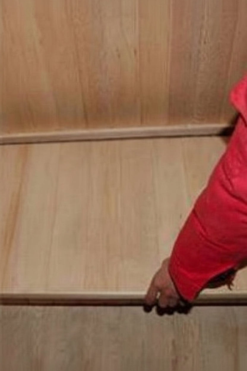 A close-up of a person's hand and red sleeve pressing down on a light-colored wooden floor expansion joint or transition strip.