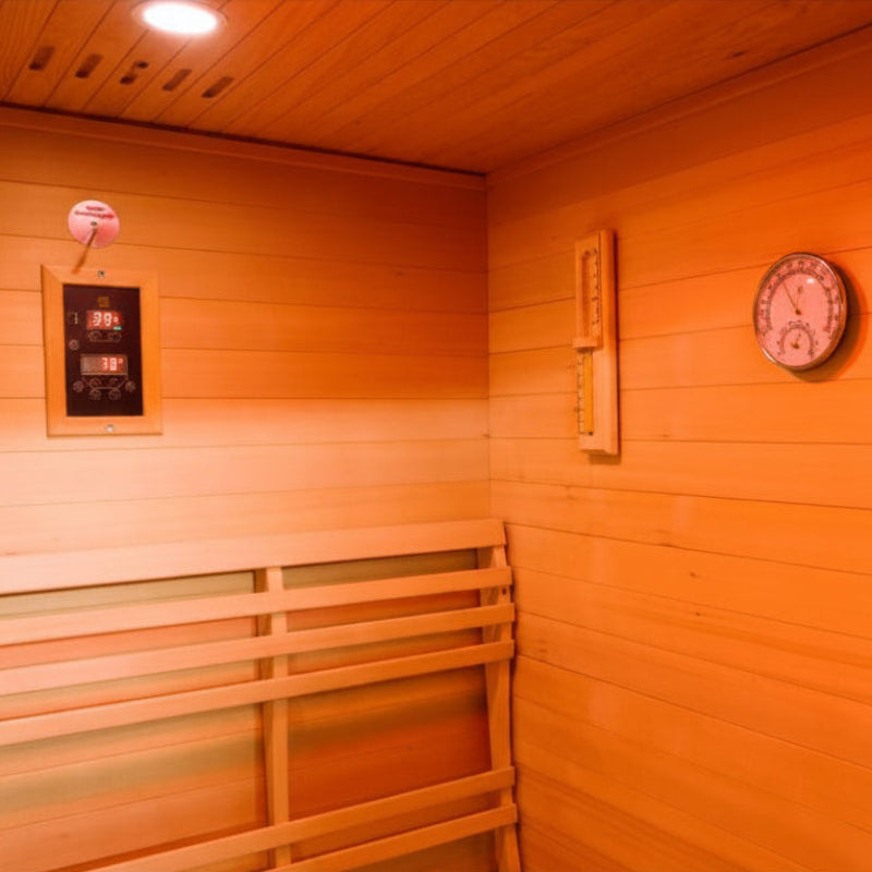 A wooden sauna interior with orange-toned wood walls and ceiling, a bench on the left, a round wall clock, and a small hanging device on the left wall.