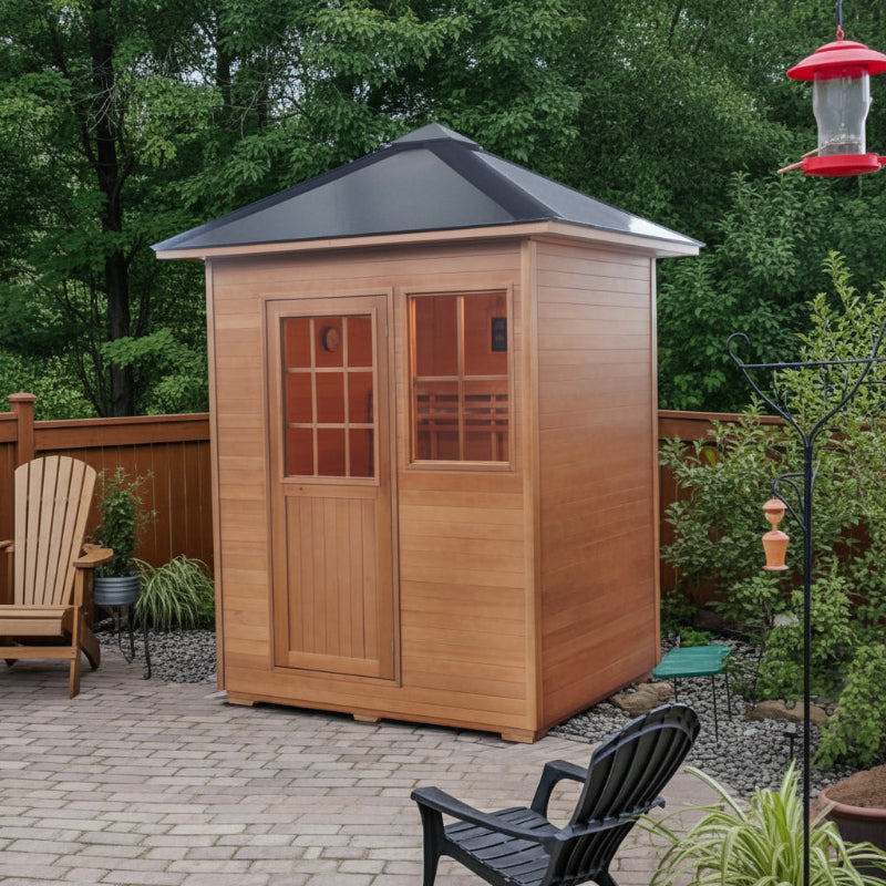 Sunray Freeport A wooden outdoor sauna with a black metal roof and glass-paneled doors, shown in a backyard setting with patio pavers, chairs, a fence, and trees.