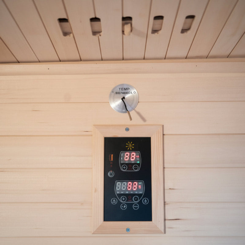 A digital sauna temperature controller with LED displays mounted on a light wood-paneled wall below a matching wood ceiling.