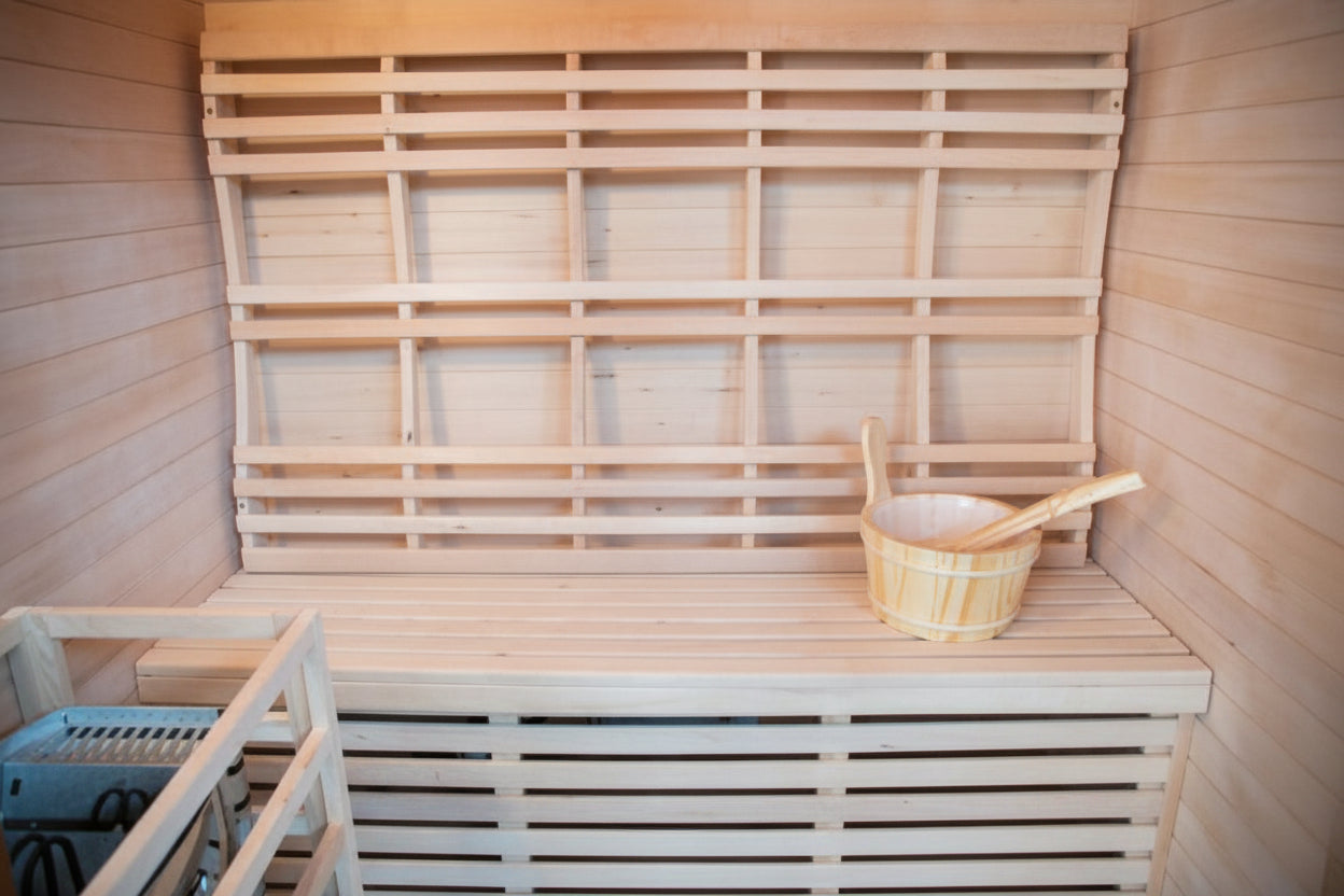 A light natural wood sauna interior with tiered wooden benches, a slatted back wall, and a woven sauna bucket with a wooden scoop resting on the front bench.