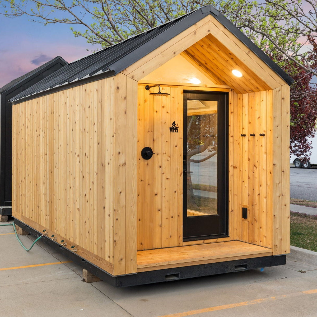 A small A-frame wooden cabin or sauna with light natural wood siding, black roof and door frame, and a glass front door, shown outdoors on a concrete surface with trees and sky in the background.