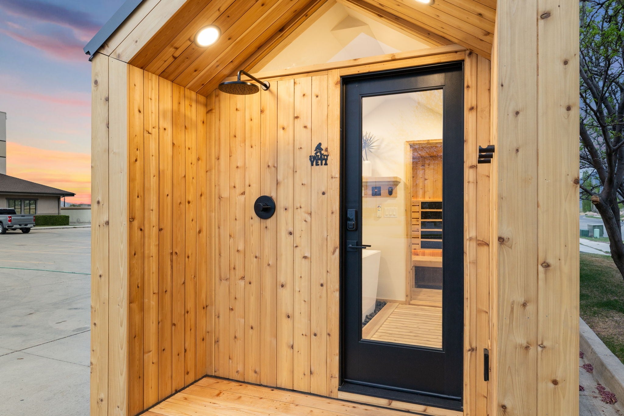 A small wooden sauna or cabin with light natural wood siding, a black-framed glass door, and a wall-mounted shower head, shown outdoors at dusk.