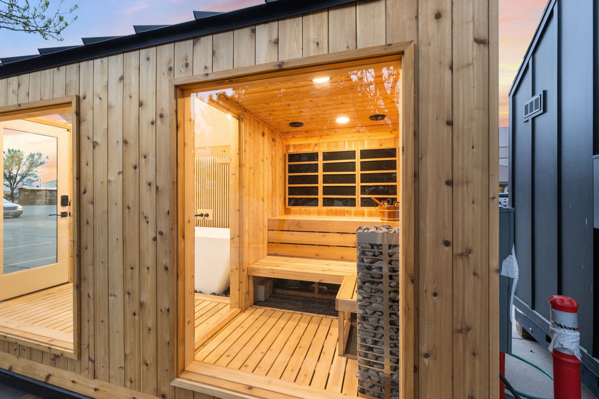 A modern wooden sauna cabin with a warm wood-paneled exterior and an open front door revealing a sauna interior with wooden benches, a stone fire column, and a glass window. A glass door is visible on the left, and another dark cabin is partially visible on the right.