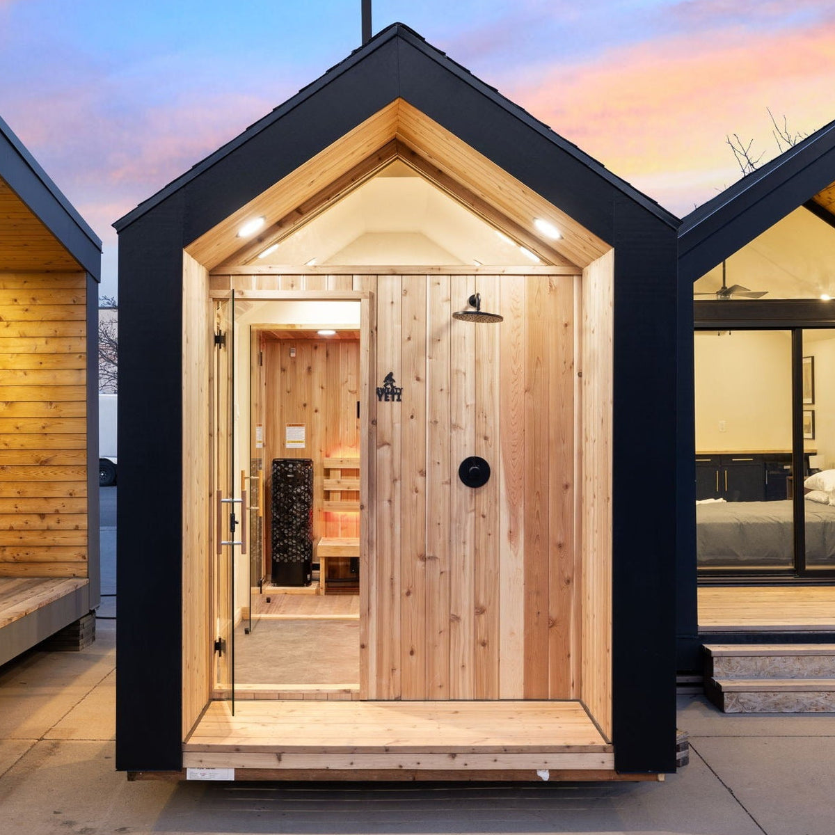 A  wooden cabin or sauna with a black exterior frame and light natural wood doors, shown open with a shower fixture installed inside. The cabin is photographed outdoors on a patio with other similar cabins and a sunset sky in the background.