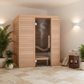 A wooden indoor sauna with a glass door and visible steam, placed in a spa-like room with tiled floor, potted plants, wall lights, and folded towels.